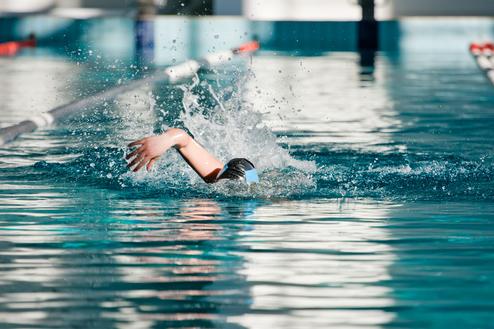 Schwimmerin in Schwarz mit blauer Badekappe zieht eine Bahn im Wasser, spritzt dabei Wasser auf.