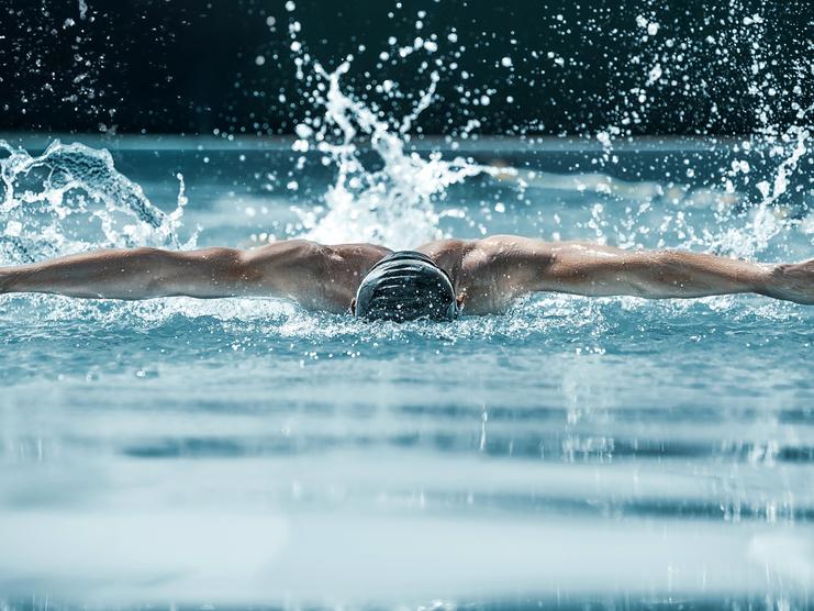 Schwimmer in einem blauen Pool macht einen Schmetterlingsschwimmzug, während Wasser spritzt und Bläschen aufsteigen.