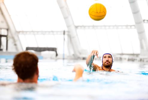 Zwei Wasserballspieler im Schwimmbecken, einer wirft einen gelben Ball, der andere schwimmt.