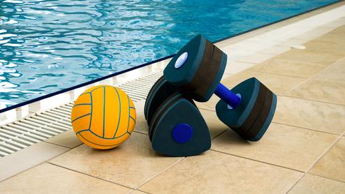 Wasserball und eine Kurzhantel liegen auf dem Boden neben einem Schwimmbecken mit blauer Wasseroberfläche.